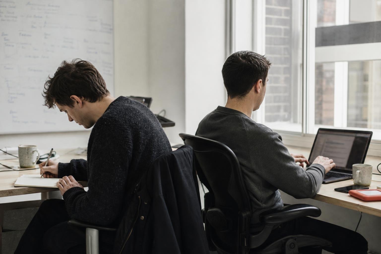 A two-person SaaS team working side by side at a window bench in a small studio
